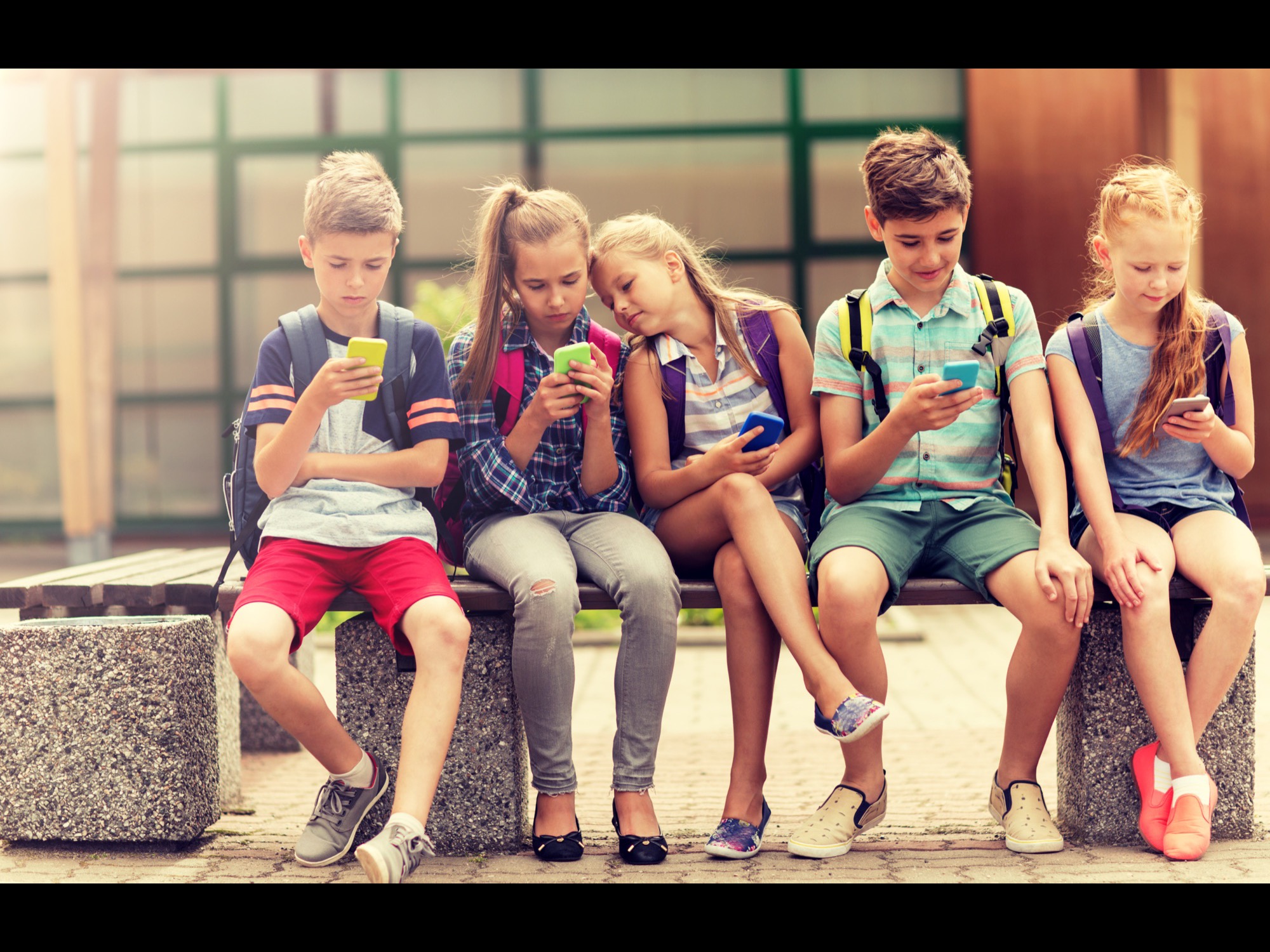 Kids sitting on a bench after school with their phones and backpacks