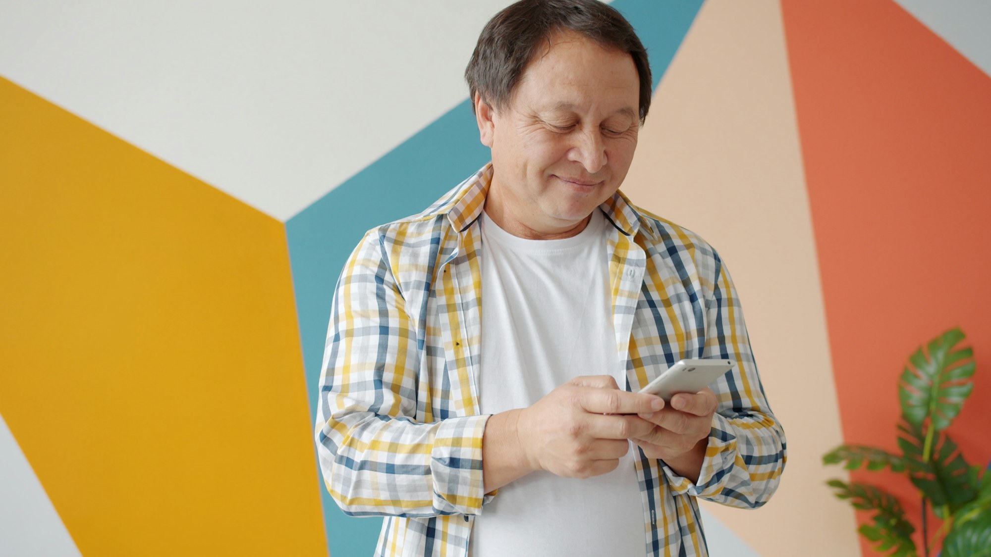 Smiling man in a checkered shirt reading his phone in front of a colorful painted wall